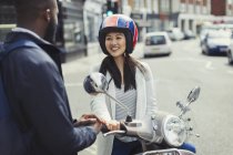 Усміхнена молода жінка на моторолері розмовляє з другом на сонячній міській вулиці — стокове фото