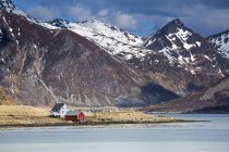 Casas remotas a lo largo del fiordo debajo de montañas escarpadas, Flakstadpollen, Lofoten, Noruega - foto de stock