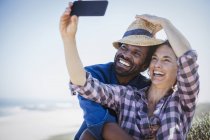 Грайлива, усміхнена багатоетнічна пара бере селфі з телефоном на сонячному літньому пляжі — стокове фото
