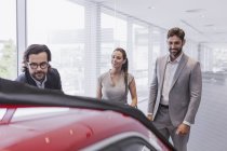 Car salesman showing new car to couple customers in car dealership showroom — Stock Photo