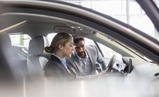 Vendedor de coches mostrando coche nuevo a la mujer en el asiento del conductor en la concesionaria de automóviles - foto de stock