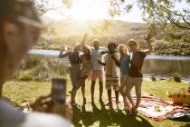 Молода жінка з телефоном фотографує друзів на сонячному літньому пікніку на березі річки — стокове фото