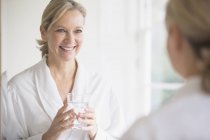 Smiling, confident mature woman in bathrobe drinking water at bathroom mirror — Stock Photo