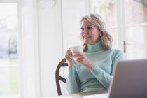 Усміхнена зріла фрілансерка п'є чай і працює на ноутбуці вдома — стокове фото