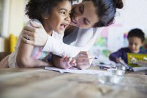 Affectionate, happy mother hugging toddler daughter coloring at table — Stock Photo