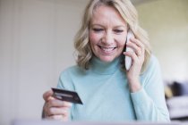 Усміхнена зріла жінка з кредитною карткою розмовляє по телефону — стокове фото