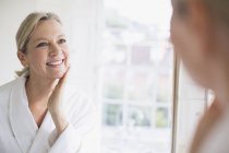 Lächelnde reife Frau berührt Gesicht im Badezimmerspiegel — Stockfoto