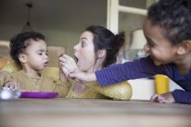 Happy caucasian mother feeding son at kitchen, multiracial family — Stock Photo