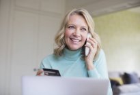 Femme mûre souriante avec carte de crédit parlant au téléphone à l'ordinateur portable — Photo de stock