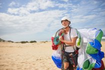 Lächelnder männlicher Gleitschirmflieger mit Ausrüstung und Fallschirm am sonnigen Strand — Stockfoto