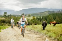 Homem de bicicleta de montanha com amigos na estrada de terra rural ao longo do pasto de vaca — Fotografia de Stock