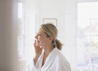 Усміхаючись зрілої жінки застосовувати зволожуючий крем на-віч у ванній кімнаті — стокове фото