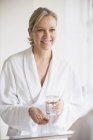 Femme mûre souriante en peignoir prenant des vitamines au miroir de salle de bain — Photo de stock