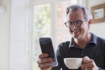 Lächelnder älterer Mann trinkt Tee und benutzt Smartphone — Stockfoto