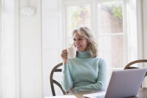 Sorrindo feminino freelancer maduro bebendo chá e trabalhando no laptop em casa — Fotografia de Stock