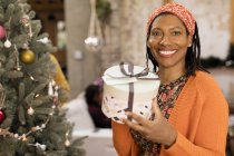 Retrato sorrindo, mulher confiante segurando presente ao lado da árvore de Natal — Fotografia de Stock