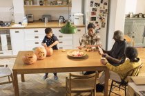 Abuelos viendo a sus nietos tallar y pintar calabazas de Halloween en la mesa de comedor - foto de stock