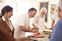 Amigos mayores viendo al chef esparciendo salsa marinara en masa de pizza en clase de cocina - foto de stock