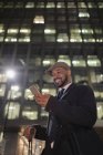 Uomo d'affari sorridente con smart phone in piedi sotto grattacielo edificio di notte — Foto stock