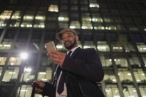 Uomo d'affari con smart phone in piedi sotto grattacielo urbano di notte — Foto stock