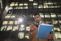 Empresário com papelada e telefone inteligente abaixo do arranha-céus urbano à noite — Fotografia de Stock