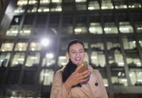 Empresária sorrindo usando telefone inteligente abaixo highrise urbano à noite — Fotografia de Stock
