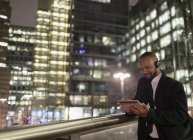 Бізнесмен з навушниками, використовуючи цифровий планшет на міському пішохідному мосту вночі — стокове фото