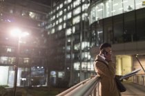 Geschäftsmann telefoniert mit Smartphone, liest nachts Papierkram auf städtischer Fußgängerbrücke — Stockfoto