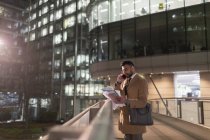 Empresario hablando por teléfono inteligente y leyendo papeleo sobre puente peatonal urbano por la noche - foto de stock