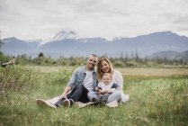Retrato pais sorridentes e bebê filho sentado no campo rural com montanhas no fundo — Fotografia de Stock
