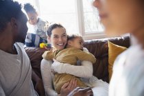 Счастливая мать обнимает маленького сына, отдыхая с семьей на диване в гостиной — стоковое фото