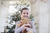 Portrait smiling, cute girl holding teddy bear in front of Christmas tree — Stock Photo