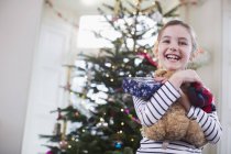 Retrato sonriente, linda chica abrazando oso de peluche delante del árbol de Navidad - foto de stock
