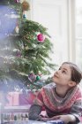 Neugieriges Mädchen mit Geschenk schaut zum Weihnachtsbaum auf — Stockfoto