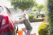 Игривая дочь распыляет мать из шланга, моет машину на солнечной дорожке — стоковое фото