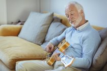 Активний старший чоловік грає на гітарі на дивані у вітальні — стокове фото