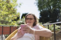 Счастливая пожилая женщина с помощью смартфона на солнечном патио — стоковое фото