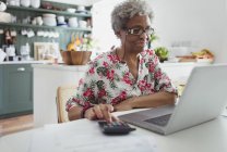 Старшая женщина оплачивает счета за ноутбук на кухне — стоковое фото