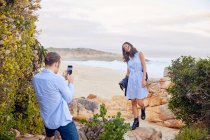 Giovane uomo con smart phone fotografare fidanzata con oceano in background — Foto stock