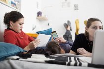 Young women friends hanging out, using smart phone, digital tablet and laptop on bed — Stock Photo