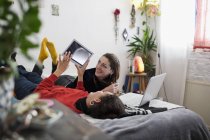 Young women friends relaxing, using digital tablet and laptop on bed — Stock Photo