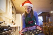 Sonriente adolescente en delantal de Navidad y Santa sombrero de hornear magdalenas en la cocina - foto de stock