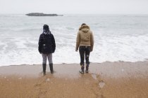 Брат и сестра в теплой одежде стоят на снежном зимнем пляже — стоковое фото