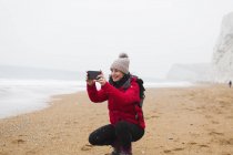 Mulher em roupas quentes usando telefone câmera na praia nevada — Fotografia de Stock