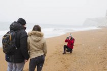 Жінка з фотоапаратом фотографує чоловіка і дочку на засніженому зимовому пляжі — стокове фото