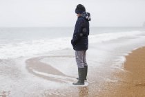 Teenage boy in rubber boots standing in winter ocean surf — Stock Photo
