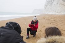 Мама с фотоаппаратом фотографирует детей на снежном зимнем пляже — стоковое фото