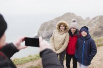 Man with camera phone photographing family in warm clothing on cliff overlooking ocean — Stock Photo