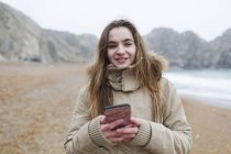 Portrait smiling teenage girl texting with smart phone on snowy winter beach — Stock Photo