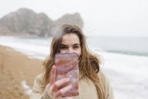 Teenage girl with camera phone taking selfie on winter ocean beach — Stock Photo